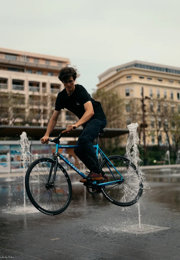 El Fakijoe riding his blue fixed gear bicycle through the fountains of a public square